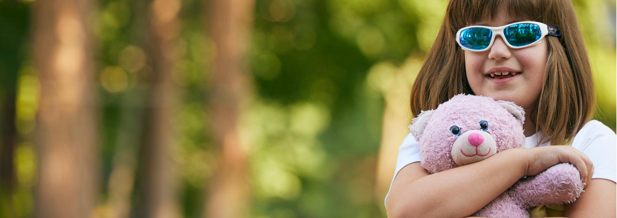 Ellamee, living with LGS, wearing sun glasses holding a pink stuffed teddy bear